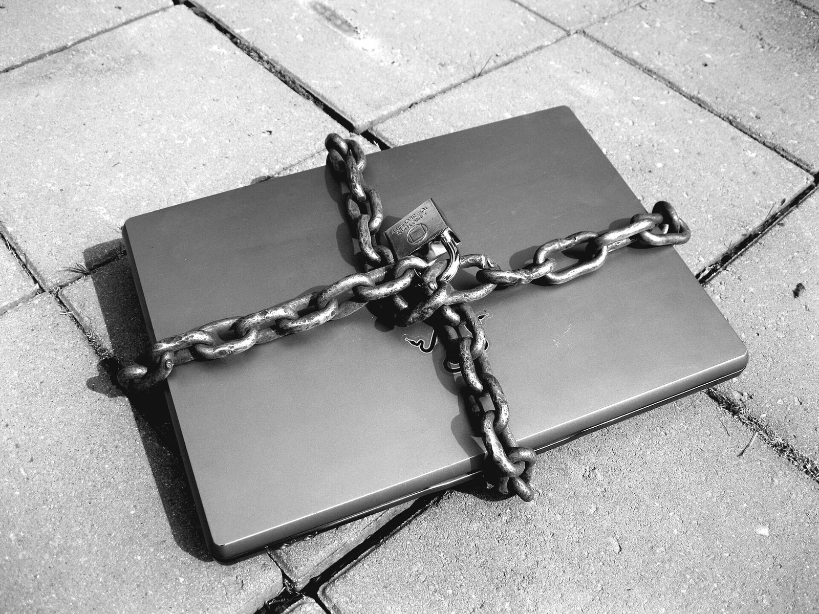A closed laptop lying on brick paving, wrapped crosswise in a heavy chain held shut with a padlock.